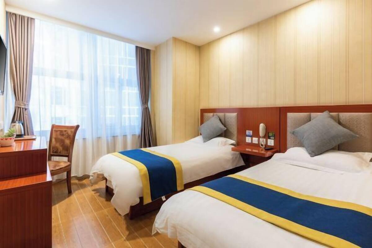 A hotel room with two beds, each with a blue and yellow bedspread, a wooden headboard, a nightstand with a lamp, and a window with curtains.