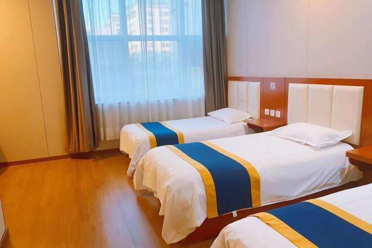 A hotel room with two beds, a wooden floor, a window with curtains, and a plain wall.