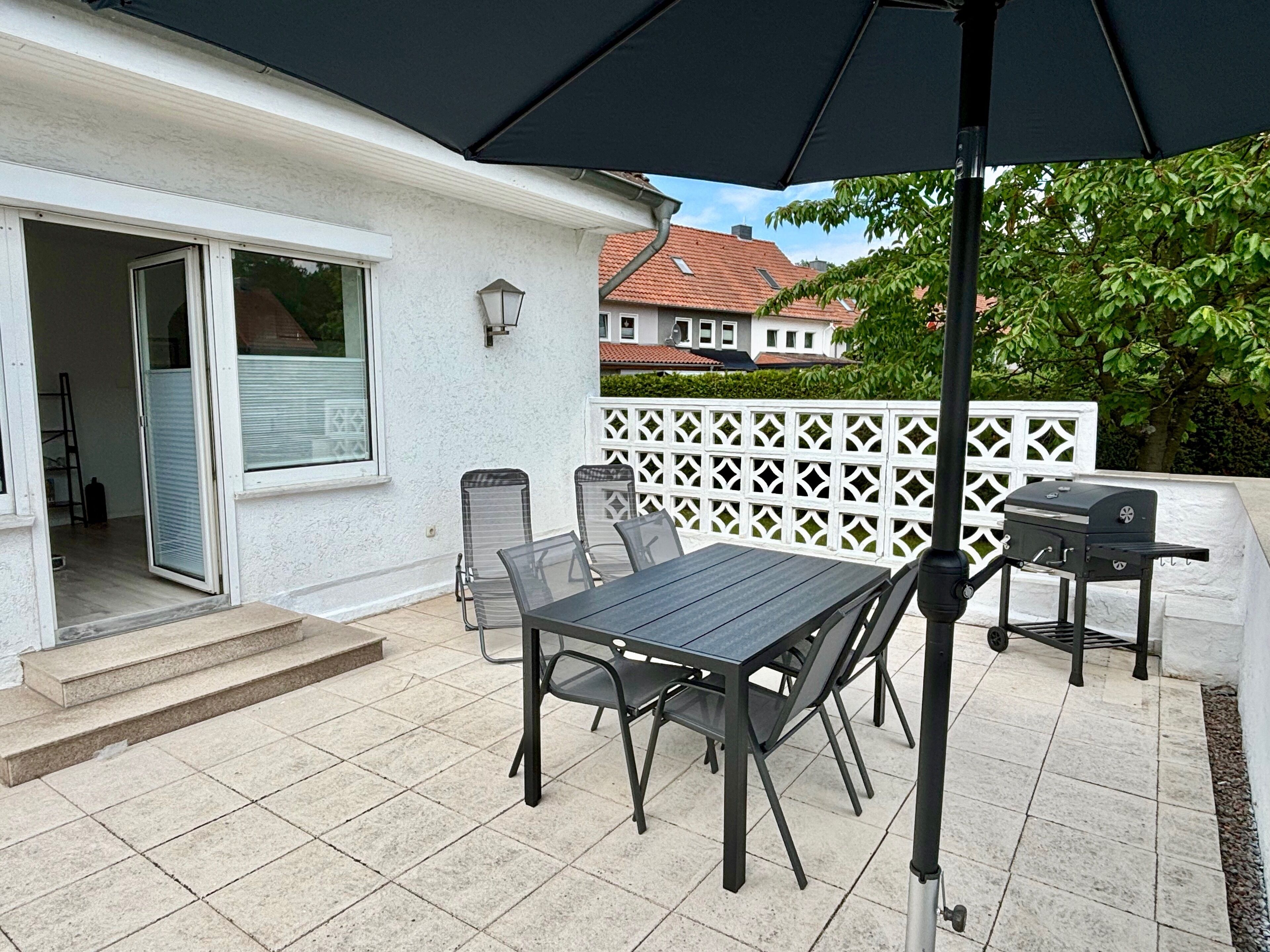 An outdoor patio with a table and chairs, a grill, and a white fence.