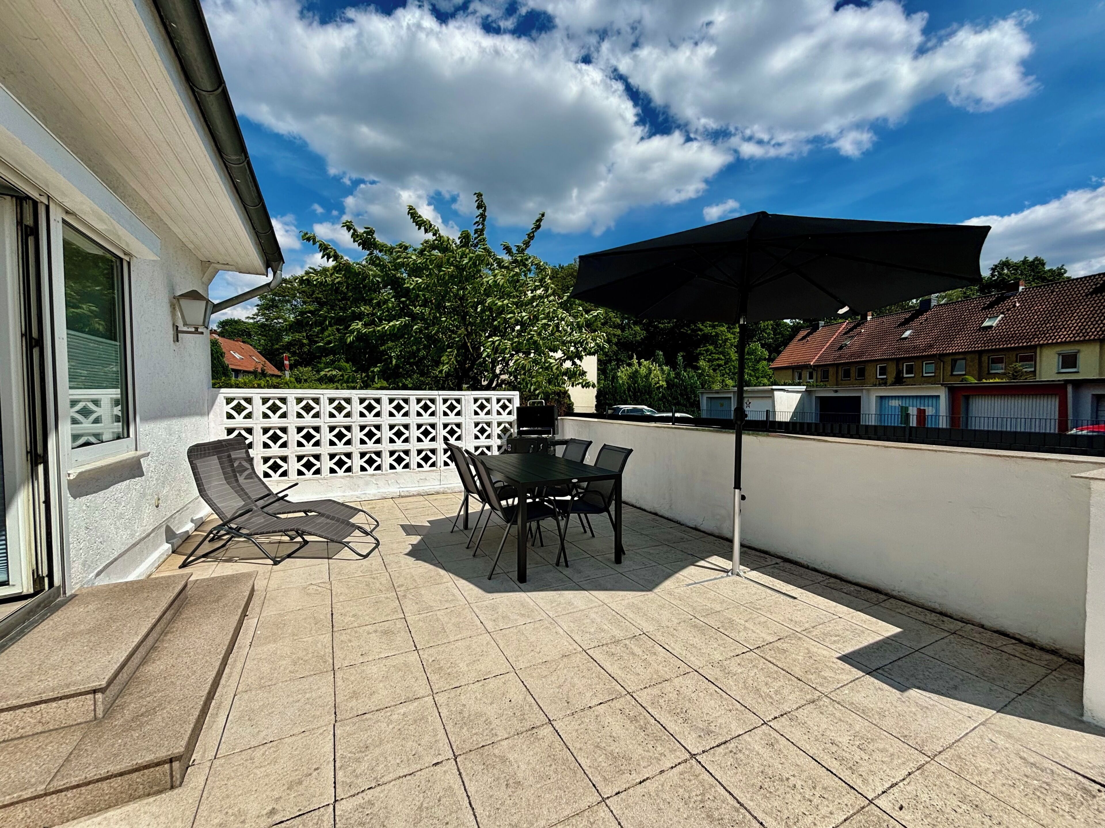 Eine Terrasse mit Tisch, Stühlen, einem schwarzen Sonnenschirm und einem weißen Zaun.