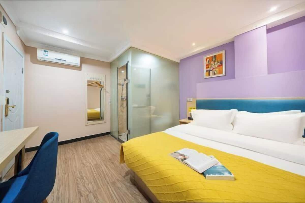 A hotel room with a bed, a desk, a chair, a mirror, and a shower.