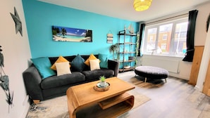 A living room with a blue wall, a black sofa with cushions, a wooden coffee table, a bookshelf, and a window with a view of buildings. - Seaside beach themed home with garden and driveway (Lytham St Annes)
