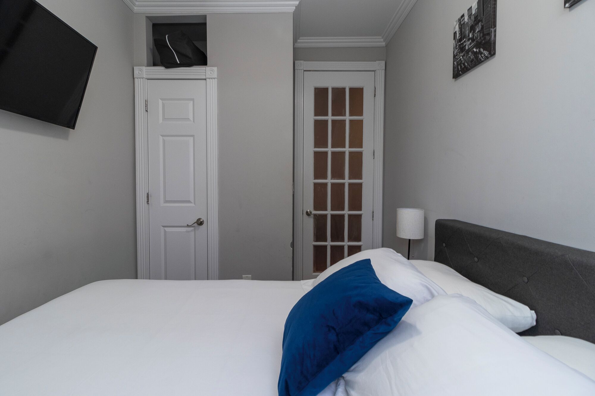 A hotel room with a bed, a door, a wall-mounted TV, and a lamp.