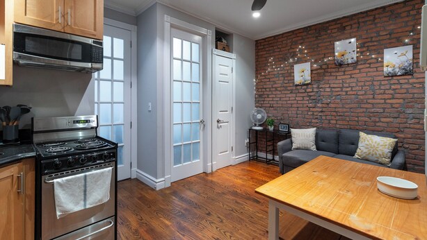 A kitchen with wooden cabinets, a stove, and a dining area with a table and a brick wall. - Lovely 2 bedroom Central Manhattan (New York)