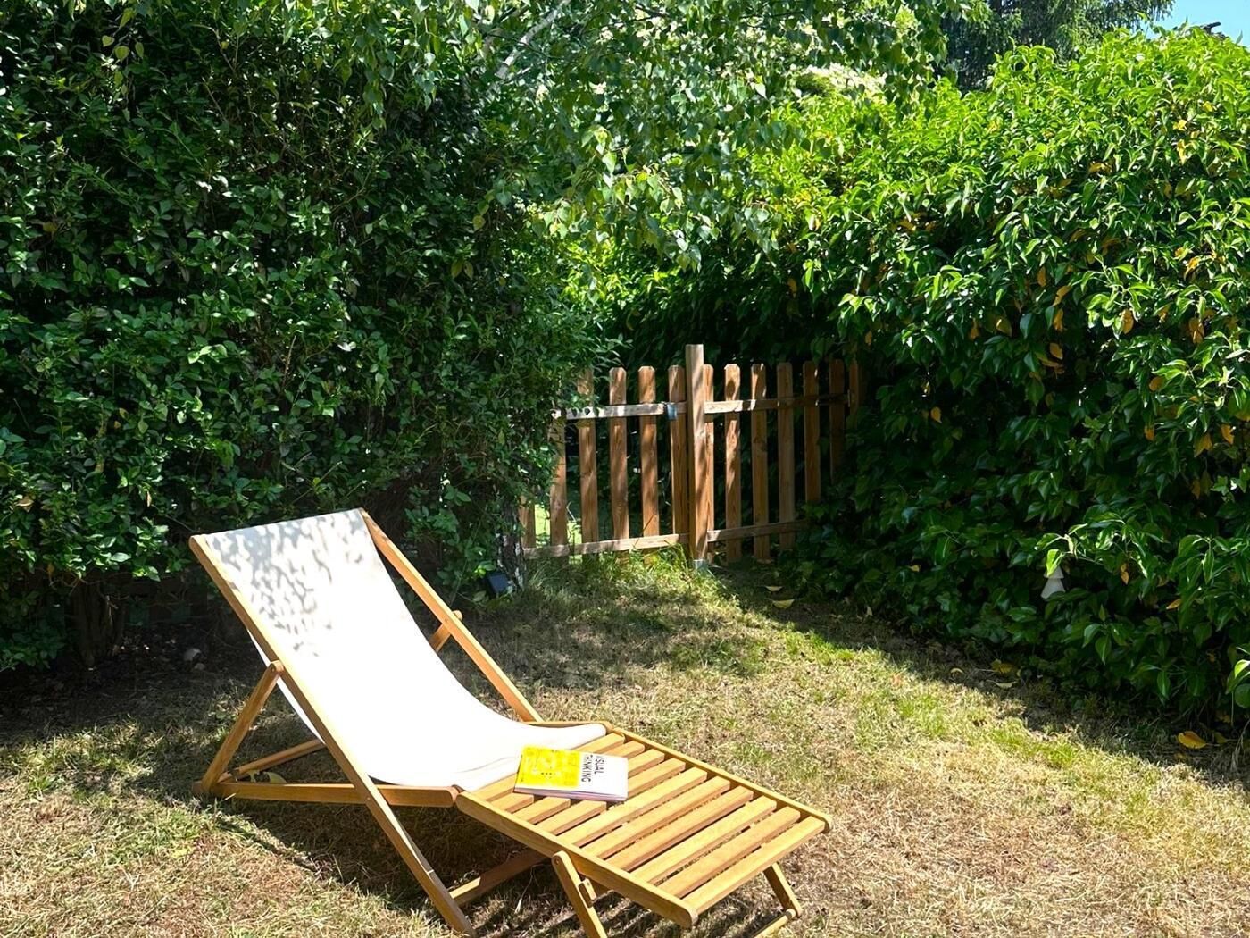 A wooden lounge chair with a book on it, placed in a grassy area with dense green foliage and a wooden fence in the background.