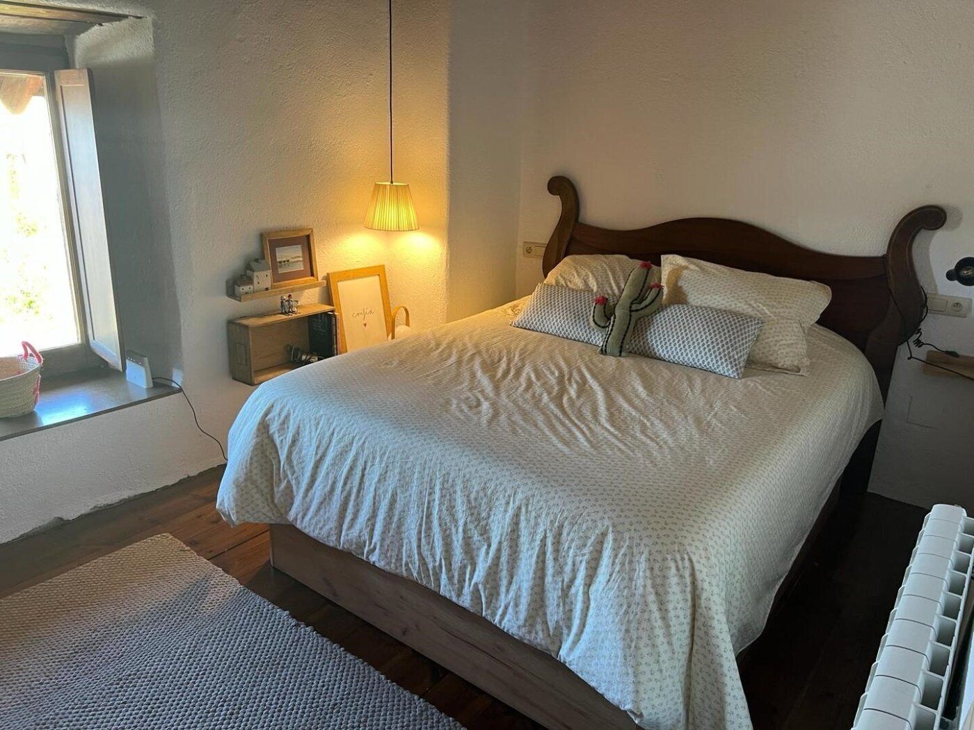 A bedroom with a wooden bed, a bedside lamp, a window with a view, and a bedside table with a lamp.