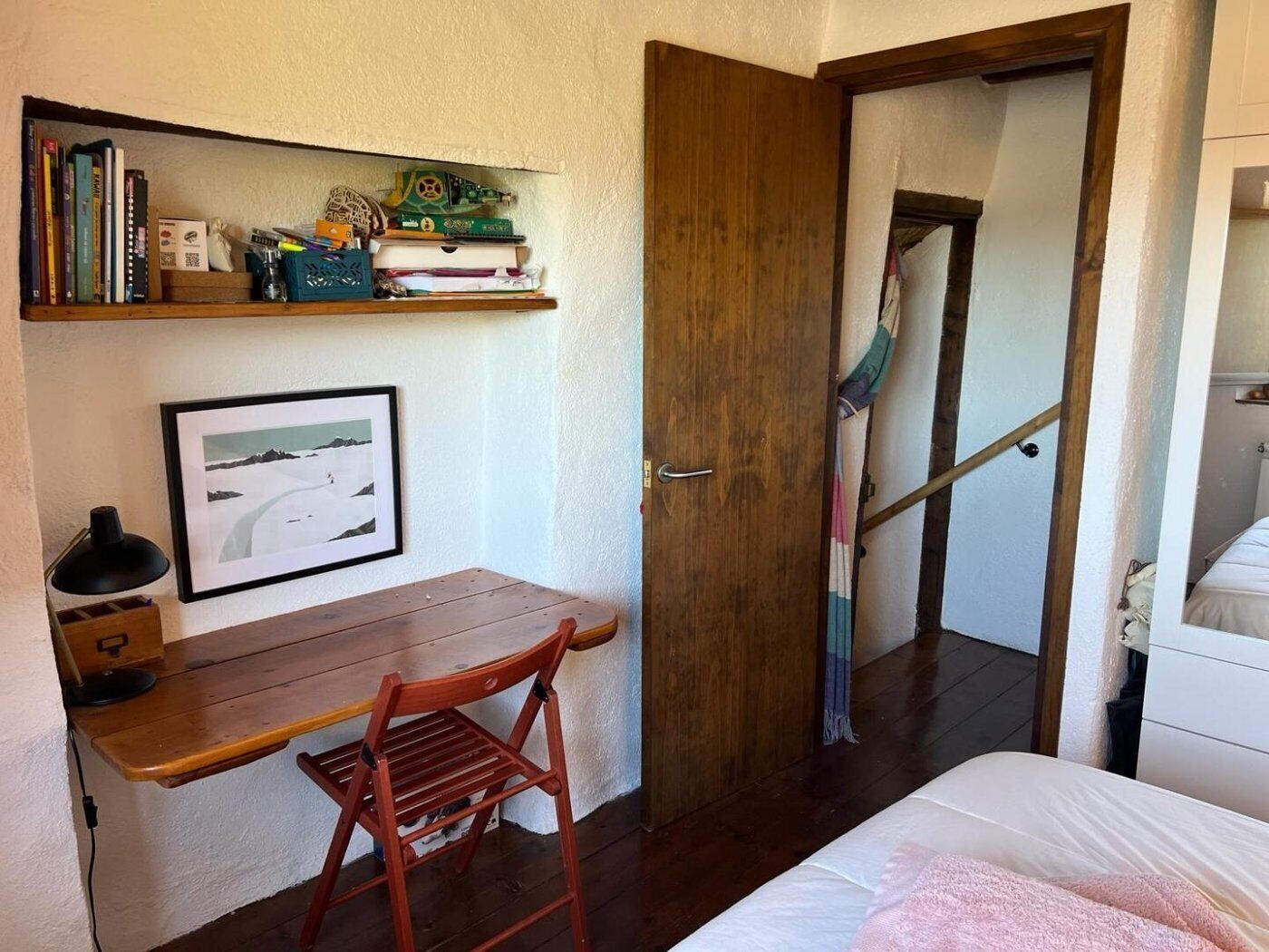 A bedroom with a wooden desk, a chair, a bed with a pink blanket, a framed picture on the wall, and a bookshelf with books and decorative items.