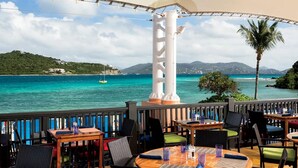 En terrasse med udendørs siddepladser med udsigt over et turkis hav, en hvid søjle og et grønt skråbjerg.