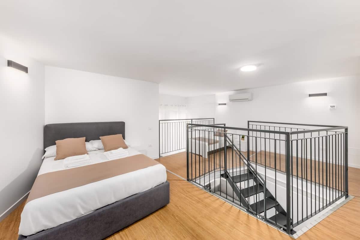 A modern bedroom with a large bed, a staircase with a railing, and a white wall with an air conditioning unit.