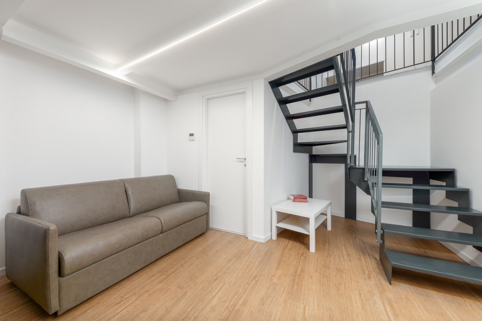 Een moderne woonkamer met een grijze bank, een witte salontafel en een trap met een zwarte balustrade.