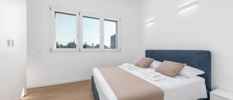 A modern bedroom with a large bed, white walls, and a wooden floor.