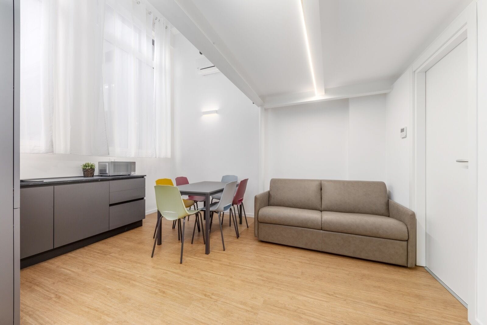 A modern living room with a beige sofa, a dining table with colorful chairs, and a kitchen area with a microwave.