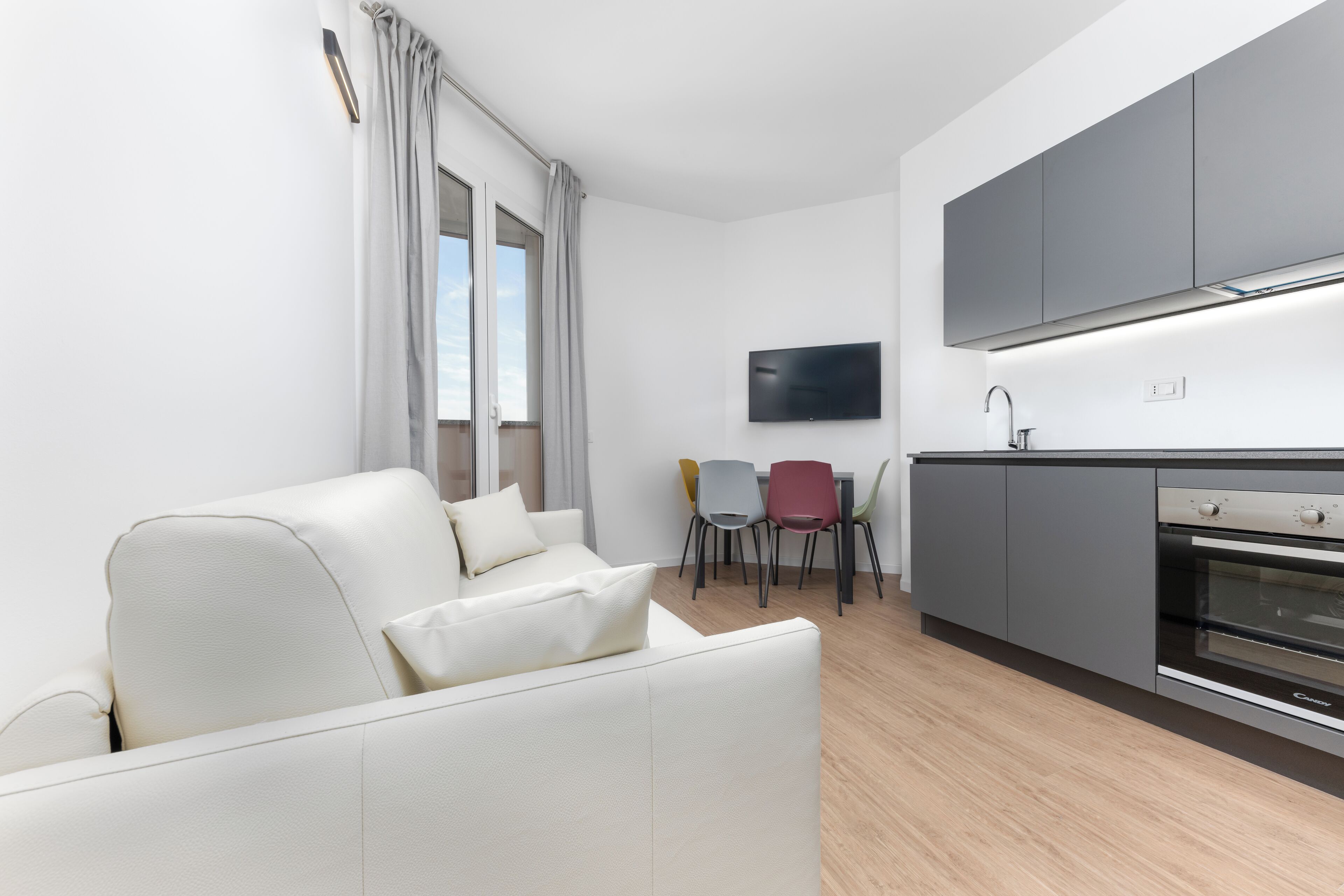 A modern living room with a white sofa, a dining area with colorful chairs, and a kitchen with grey cabinets and a built-in oven.