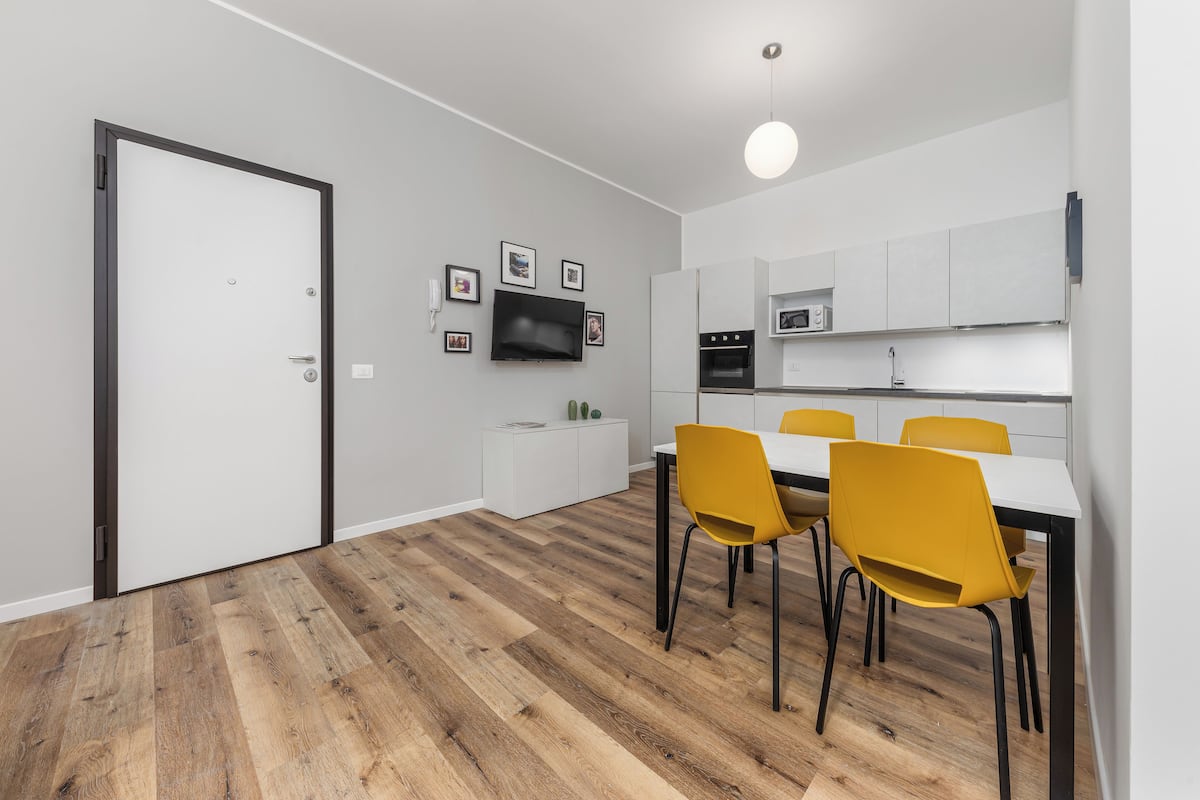A modern kitchen with white cabinets, a built-in oven, and a microwave. There is a dining table with four yellow chairs.