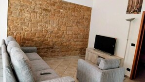 Un salon avec un mur en pierre, des canapés gris, un téléviseur à écran plat posé sur un meuble en bois et un lampadaire.