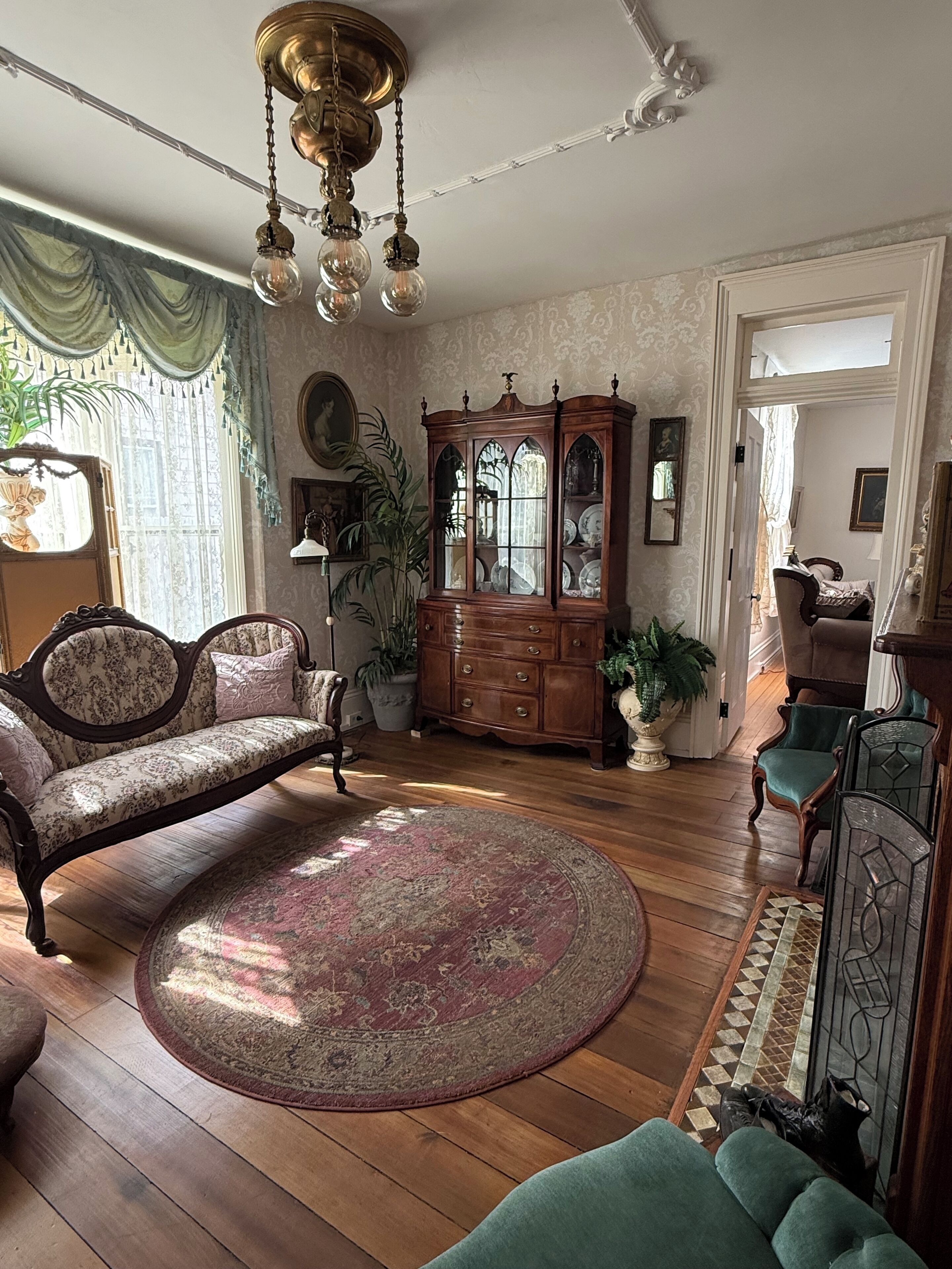 Un salon traditionnel avec un plancher en bois, un tapis à motifs, un fauteuil vert, un meuble en bois doté de portes en verre et un luminaire au plafond.