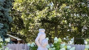 Estátua branca ao ar livre de uma mulher em pedestal, com base de fonte, rodeada por flores brancas e cerca de madeira.