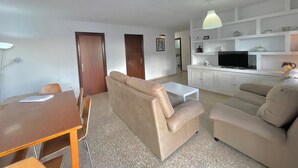 A living room with a sofa, a TV, and a small table. There are two doors and a kitchen area in the background.