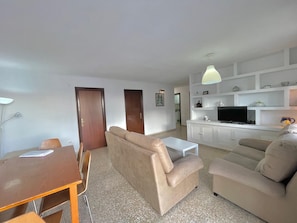 A living room with a sofa, a TV, and a small table. There are two doors and a kitchen area in the background.