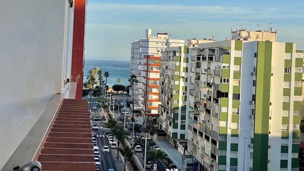A high-rise building with multiple balconies, a view of the sea, and a busy street with cars and palm trees.