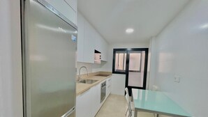 A compact kitchen with a stainless steel refrigerator, a sink, and a dining area with a glass table and chairs. - TERRASOL AZUCARERA 4 (Torre del Mar)
