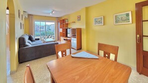 Una sala de estar con un sofá, una mesa de comedor y sillas. Hay un mueble para televisión con un televisor y un armario. La habitación tiene paredes amarillas y una ventana con vista a edificios.