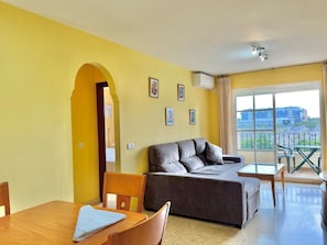 A room with a sofa, a dining table, a chair, a balcony with a view of buildings, and a ceiling fan.