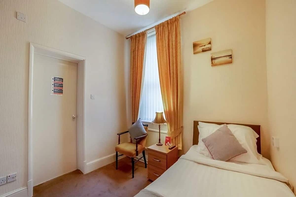 A hotel room with a bed, a chair, a nightstand, a lamp, and a window with curtains.