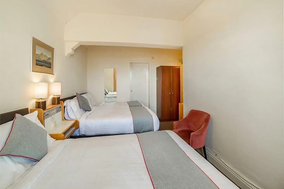 A hotel room with two beds, a wooden wardrobe, a red chair, and a bedside table with lamps.