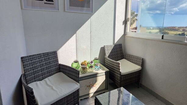 Een balkon met rieten meubilair, een kleine tafel en decoratieve objecten. Twee kunstwerken hangen in lijsten aan de muur.