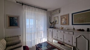 Een woonkamer met een bank, een glazen salontafel, een witte kast met decoratieve objecten, en een raam met transparante gordijnen.