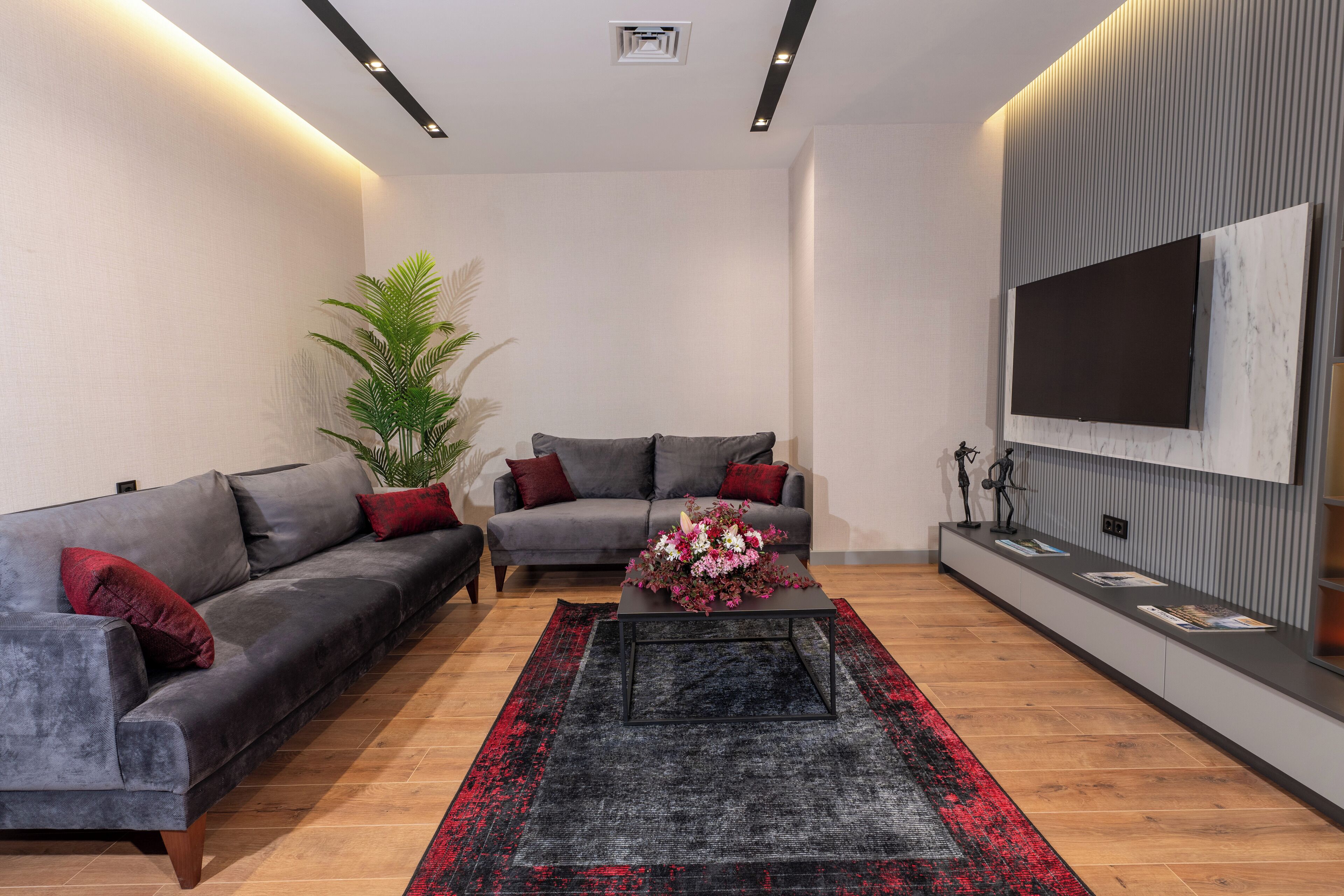 A modern living room with two grey sofas, a black coffee table with flowers, a large potted plant, and a mounted TV on the wall.