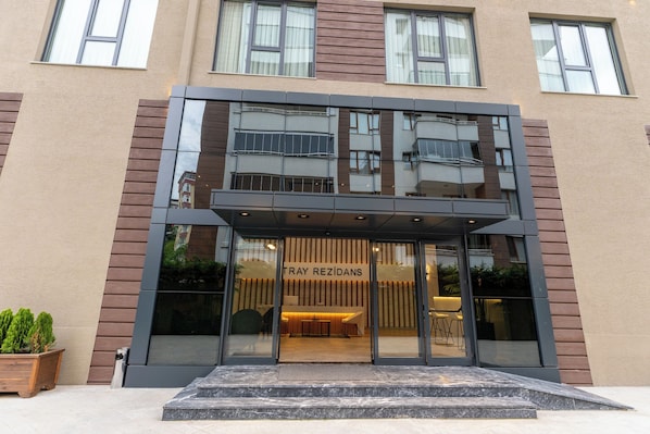 Un edificio moderno con entrada de vidrio, un cartel que dice "TRAY REZIDANS" y un pequeño espacio al aire libre con plantas.