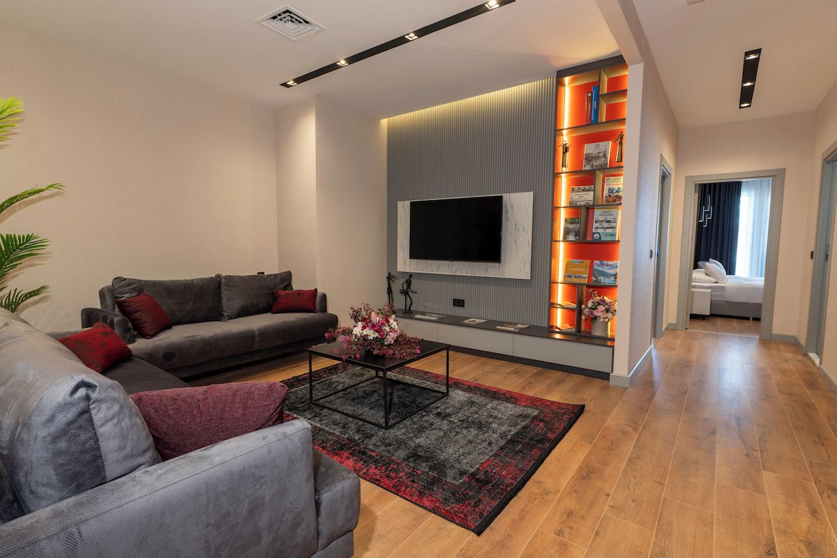 A modern living room with a grey sofa, a black coffee table, and a TV mounted on the wall. There is a bookshelf with books and decorative items, and a red and black rug on the floor.