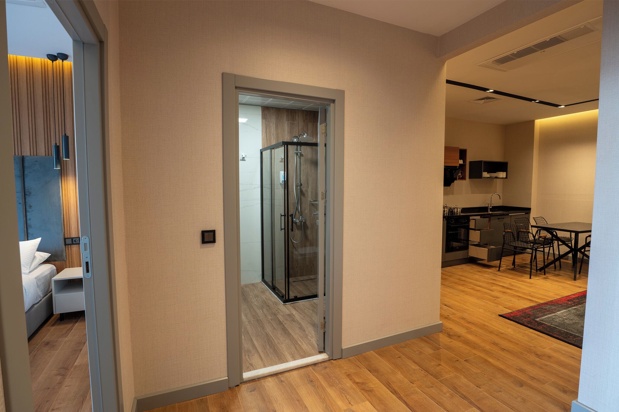 Una habitación de hotel moderna con piso de madera, cabina de ducha de vidrio, zona de comedor con mesa y sillas, y área de cocina con gabinetes y microondas.