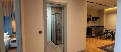 Una habitación de hotel moderna con piso de madera, cabina de ducha de vidrio, zona de comedor con mesa y sillas, y área de cocina con gabinetes y microondas.