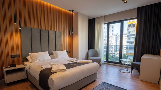 A modern hotel room with a large bed, a desk, a chair, and a view of the city through large windows. - TRAY TRABZON  (Yomra)