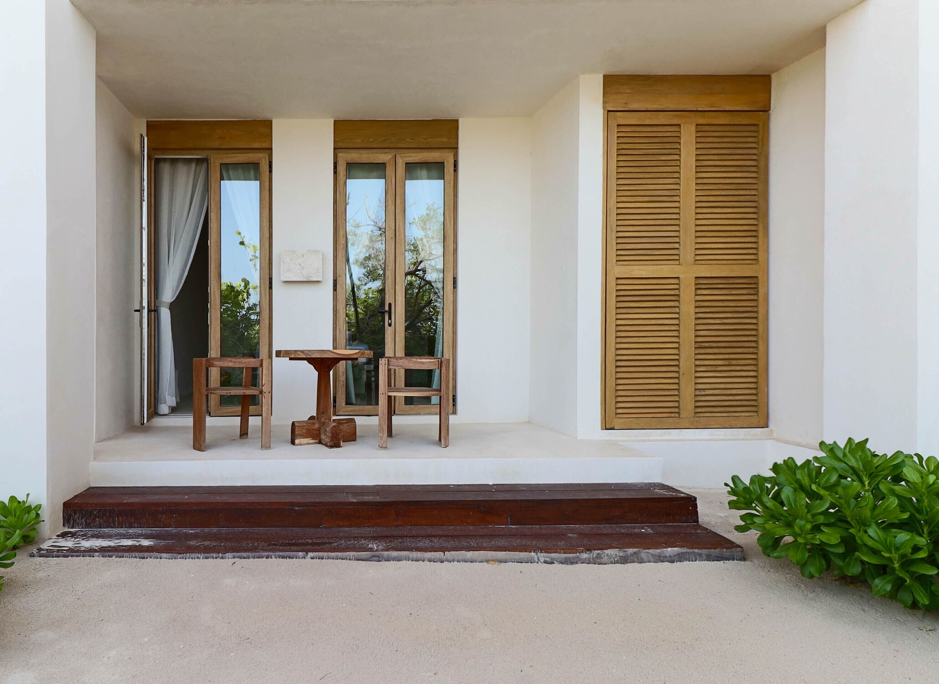A covered outdoor seating area with a table and two chairs, a wooden door, and a window with shutters.