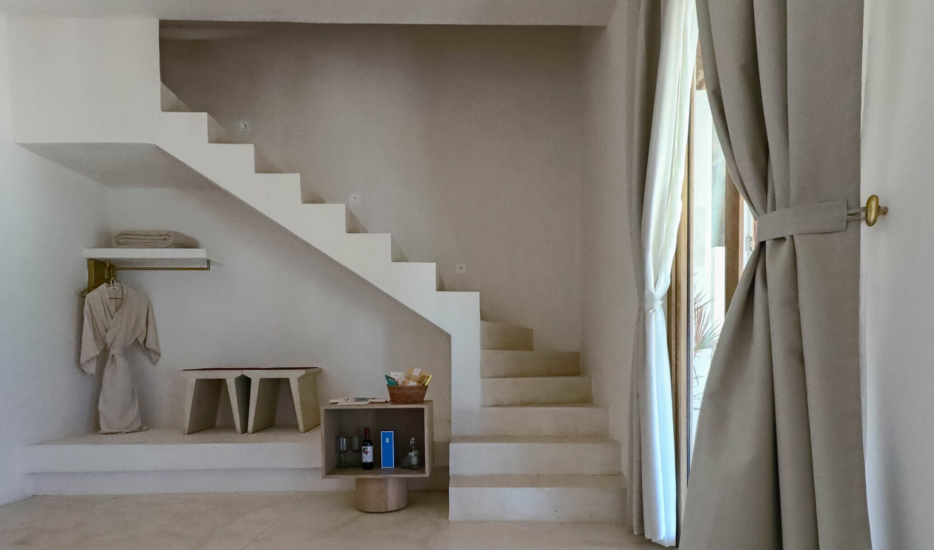 A staircase with a minimalist design, a robe hanging on a rack, a small shelf with a basket, and a door with curtains.