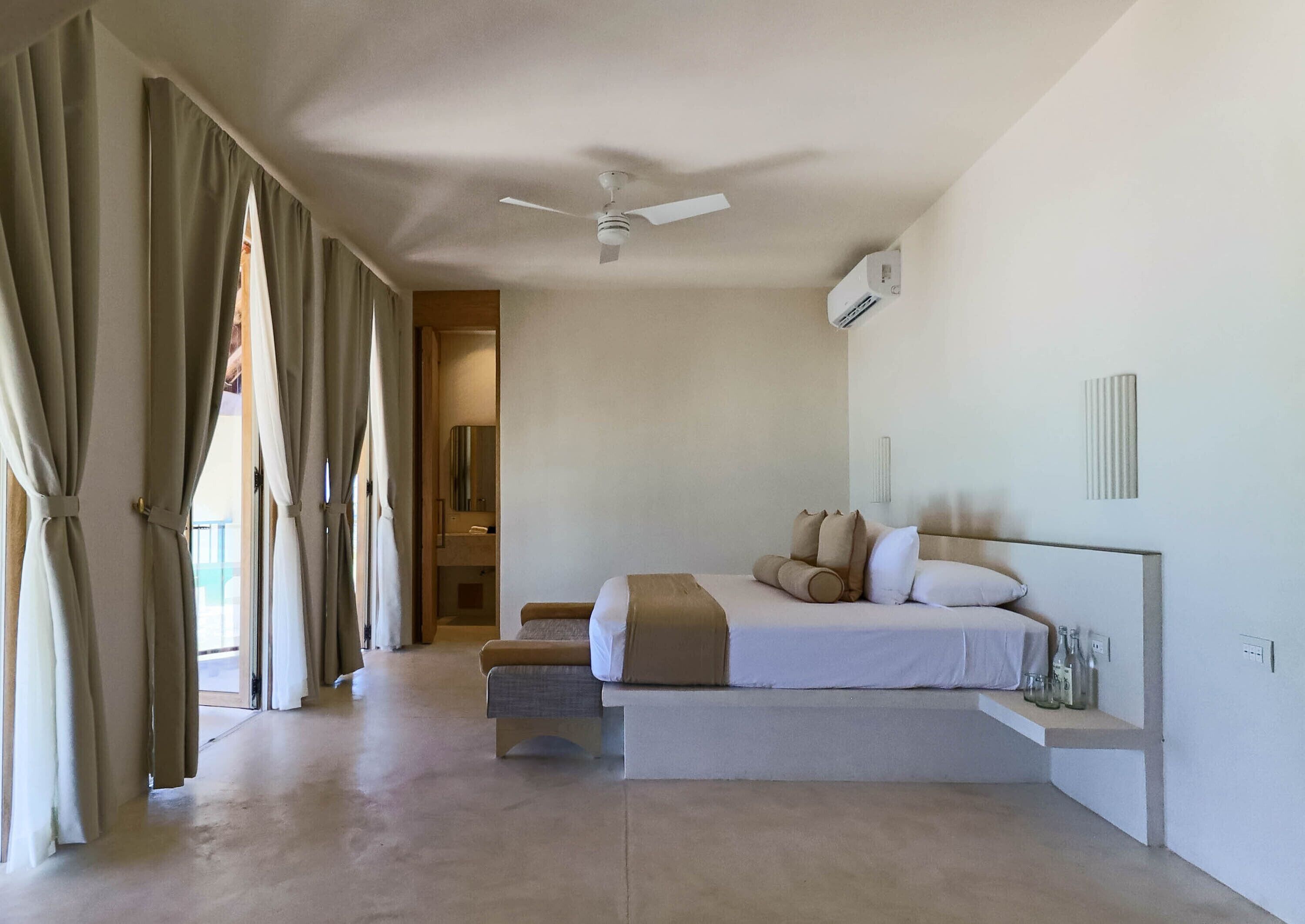 A bedroom with a bed, a ceiling fan, an air conditioning unit, and a bathroom visible through an open door.