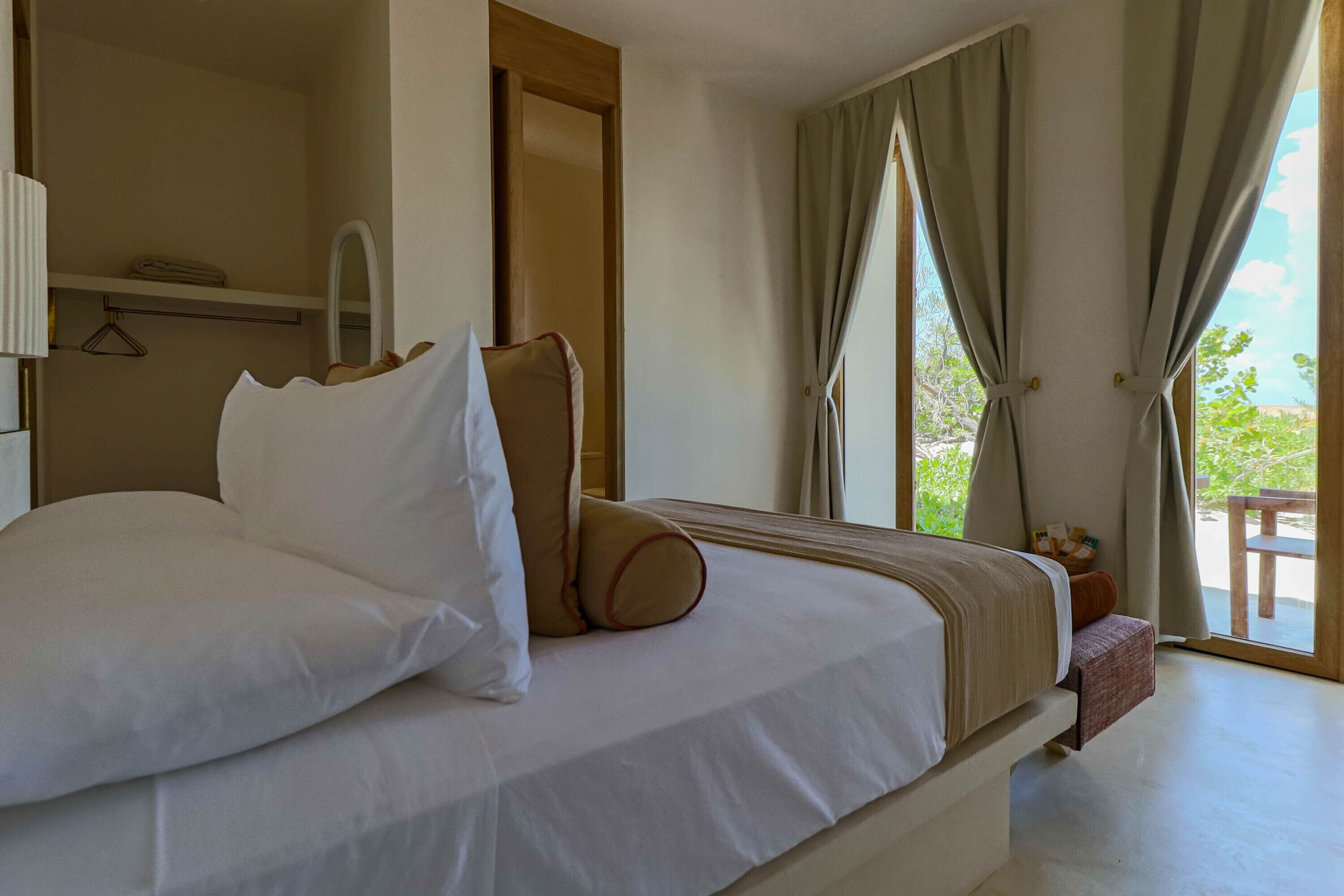 A neatly made bed with pillows and a blanket, a bedside table with a lamp, a mirror, and a view of the outdoors through a window with curtains.