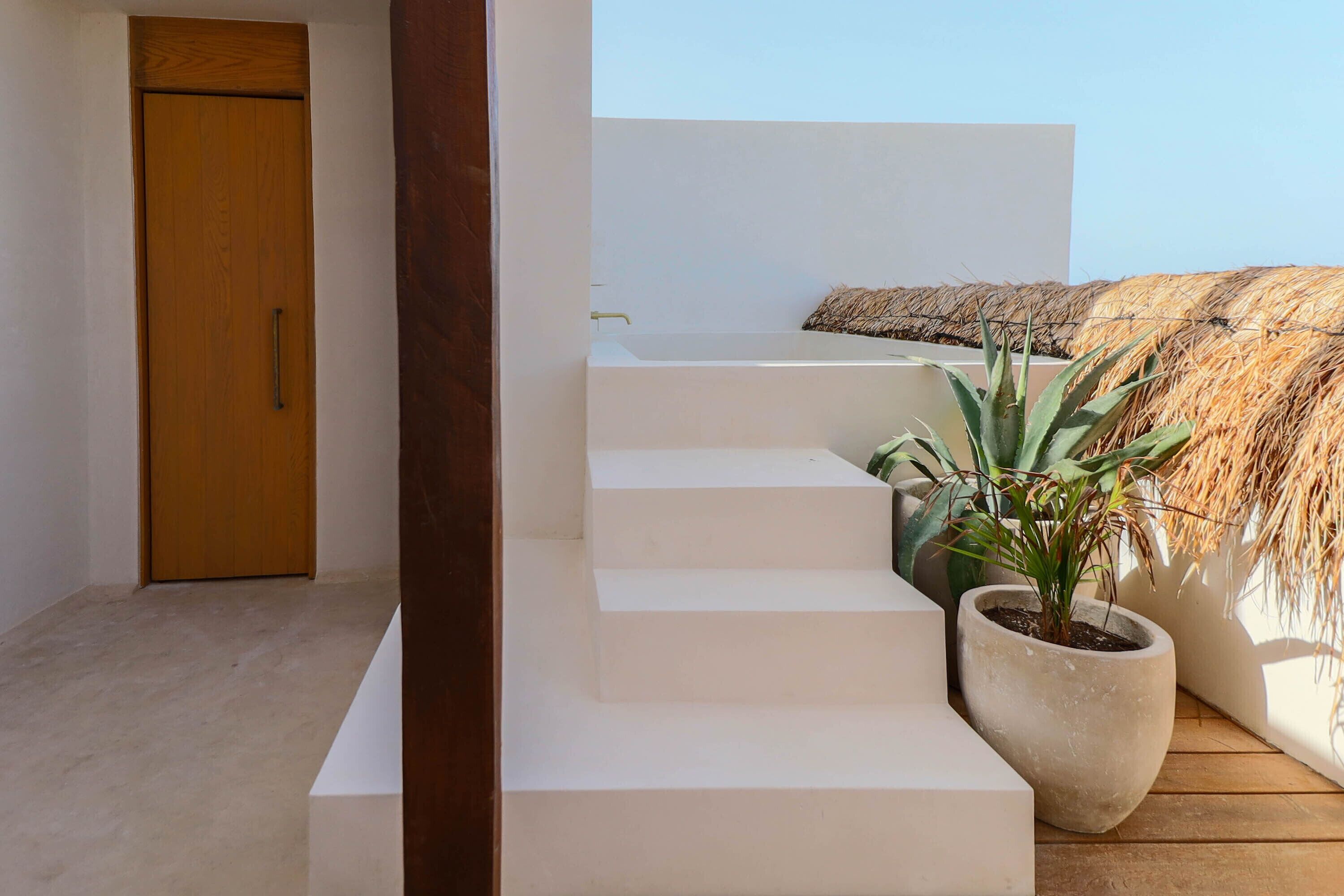 A modern staircase leading to a white room with a wooden door, a potted plant, and a thatched roof structure.