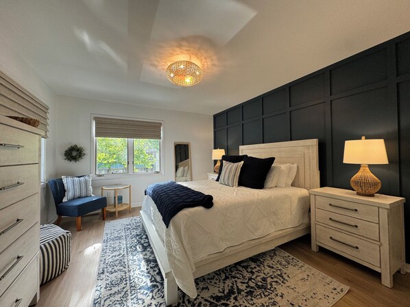 A bedroom with a large bed, bedside tables, a dresser, a window with blinds, a ceiling light, and a view of greenery outside.