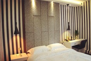 A hotel room with a bed, a desk, and a chair. There is a wall-mounted lamp and a small table with a plant.