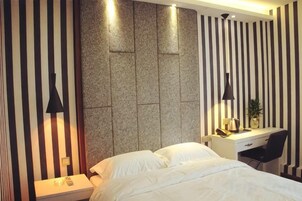 Habitación de hotel con cama, escritorio y silla. Se observa una lámpara de pared y una pequeña mesa con una planta.