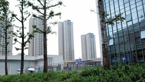 Un edificio moderno con fachada de vidrio, rodeado de vegetación y otros edificios altos al fondo.