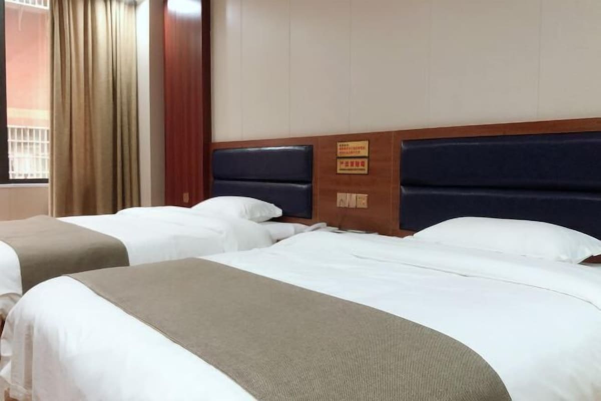 A hotel room with two beds, beige bedding, a dark headboard, and a window with curtains.