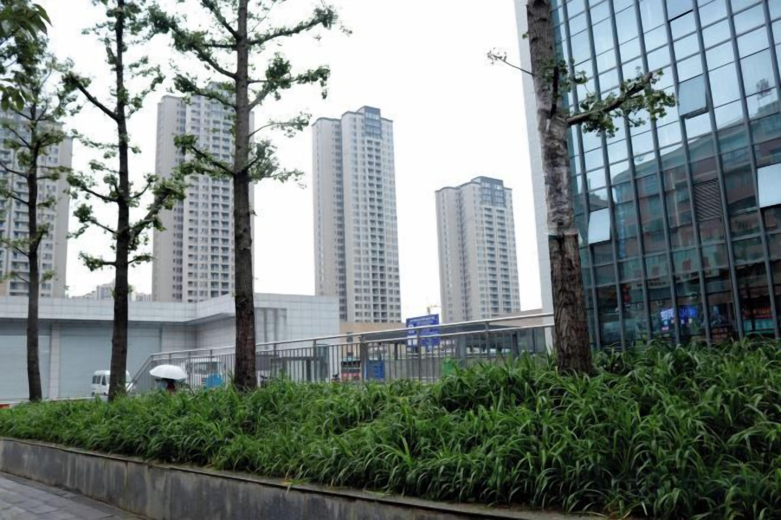 Edificio moderno con fachada de vidrio, rodeado de vegetación y junto a edificios residenciales de gran altura.