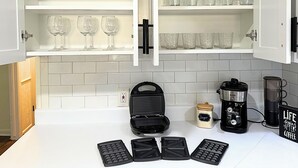Een keuken met een wit aanrecht, een wafelijzer, een koffiezetapparaat en glaswerk op planken.