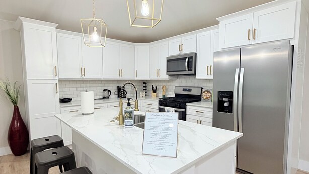 A modern kitchen with white cabinets, a large island with bar stools, a stainless steel refrigerator, and pendant lighting. - Modern 4BR/2B Home, with HotTub, Near DFW Airport (Irving)